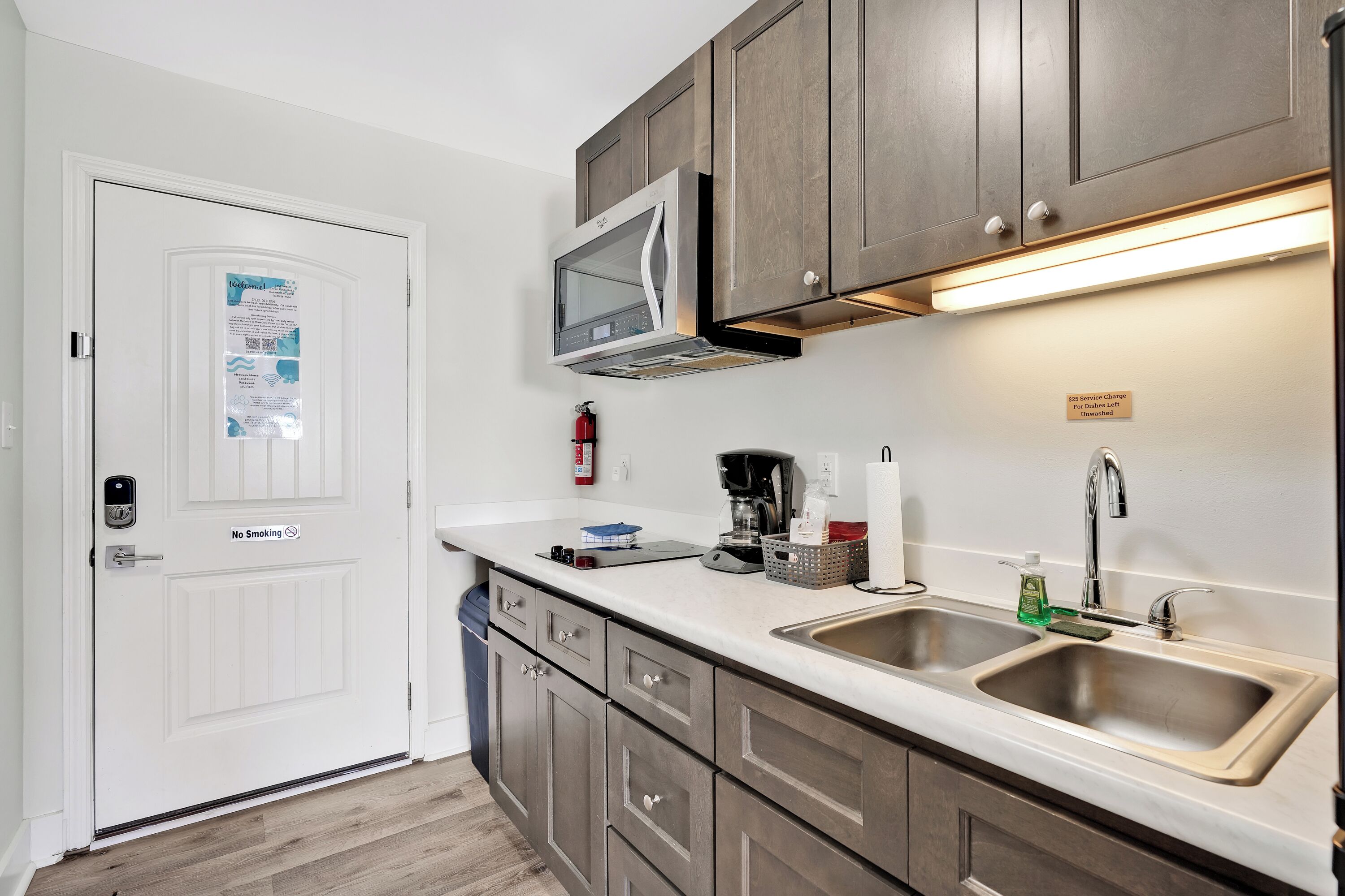 A kitchen with a microwave, double sink, and cabinets. There is a sign on the wall and a fire extinguisher.