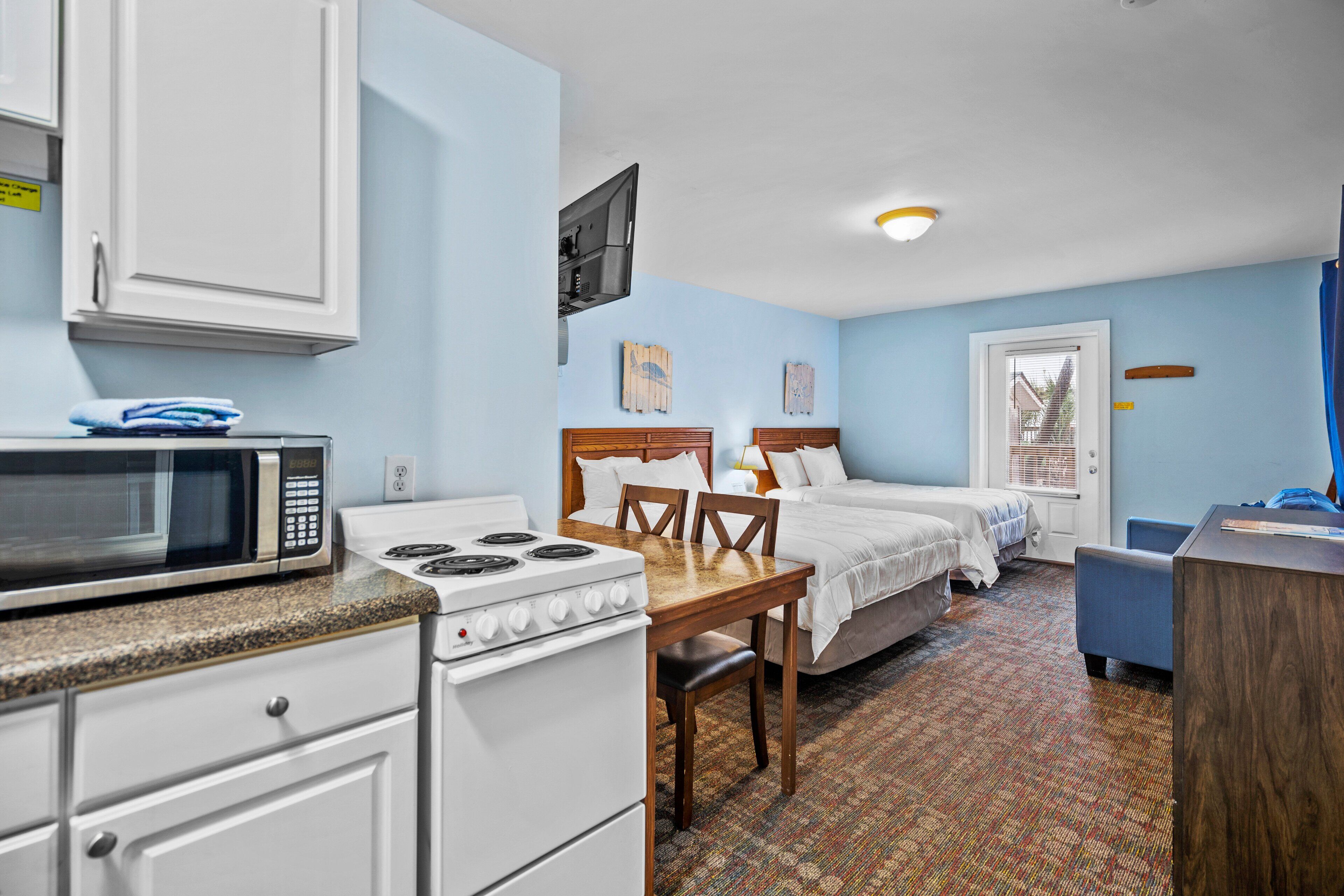 A compact hotel room with a kitchenette, a bed with two pillows, a desk, and a window with blinds.