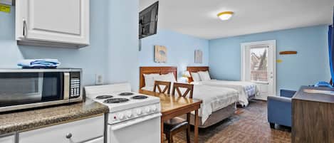 A compact hotel room with a kitchenette, a bed with two pillows, a desk, and a window with blinds.