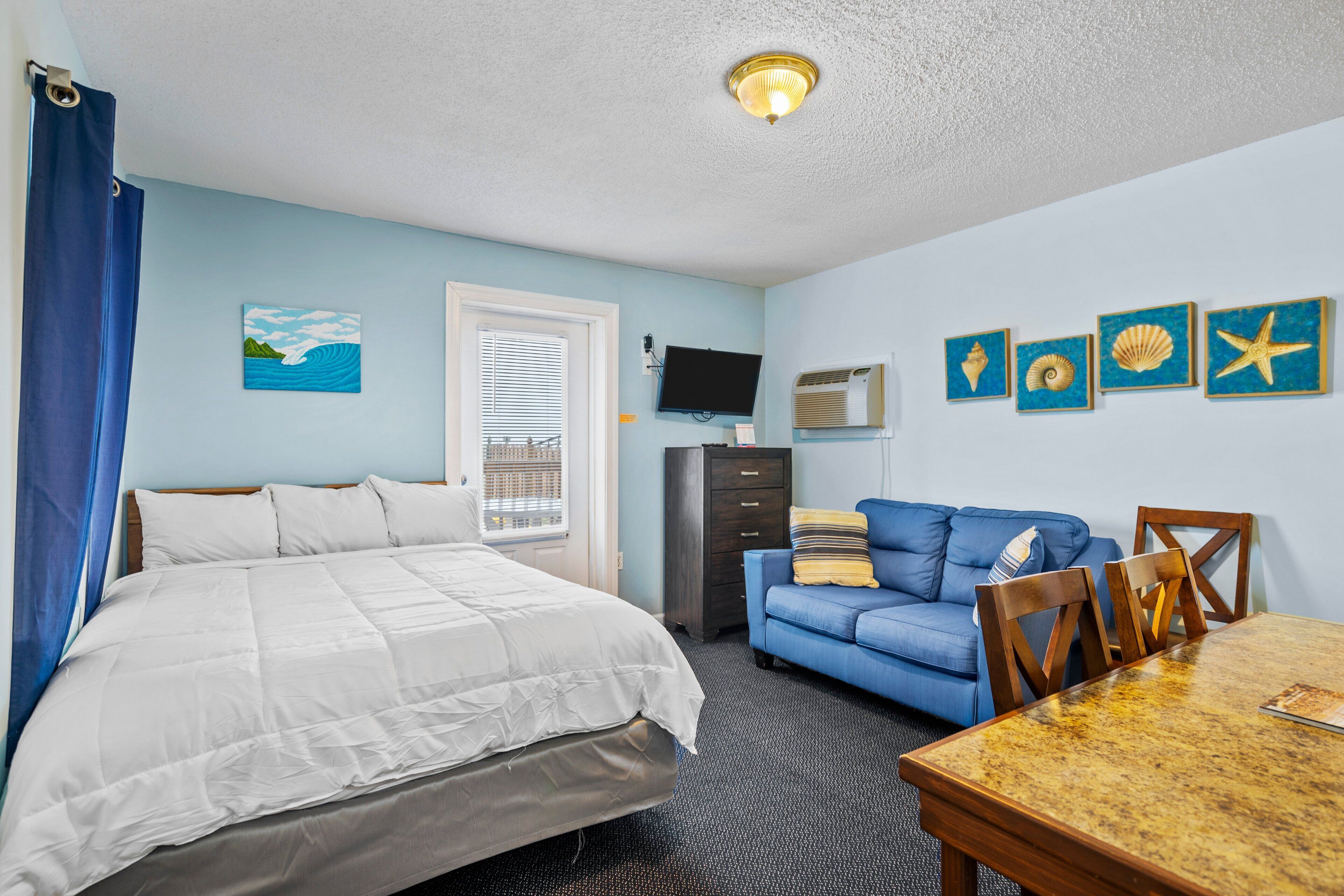 A hotel room with a bed, a blue sofa, a wooden dining table, and wall-mounted artwork.