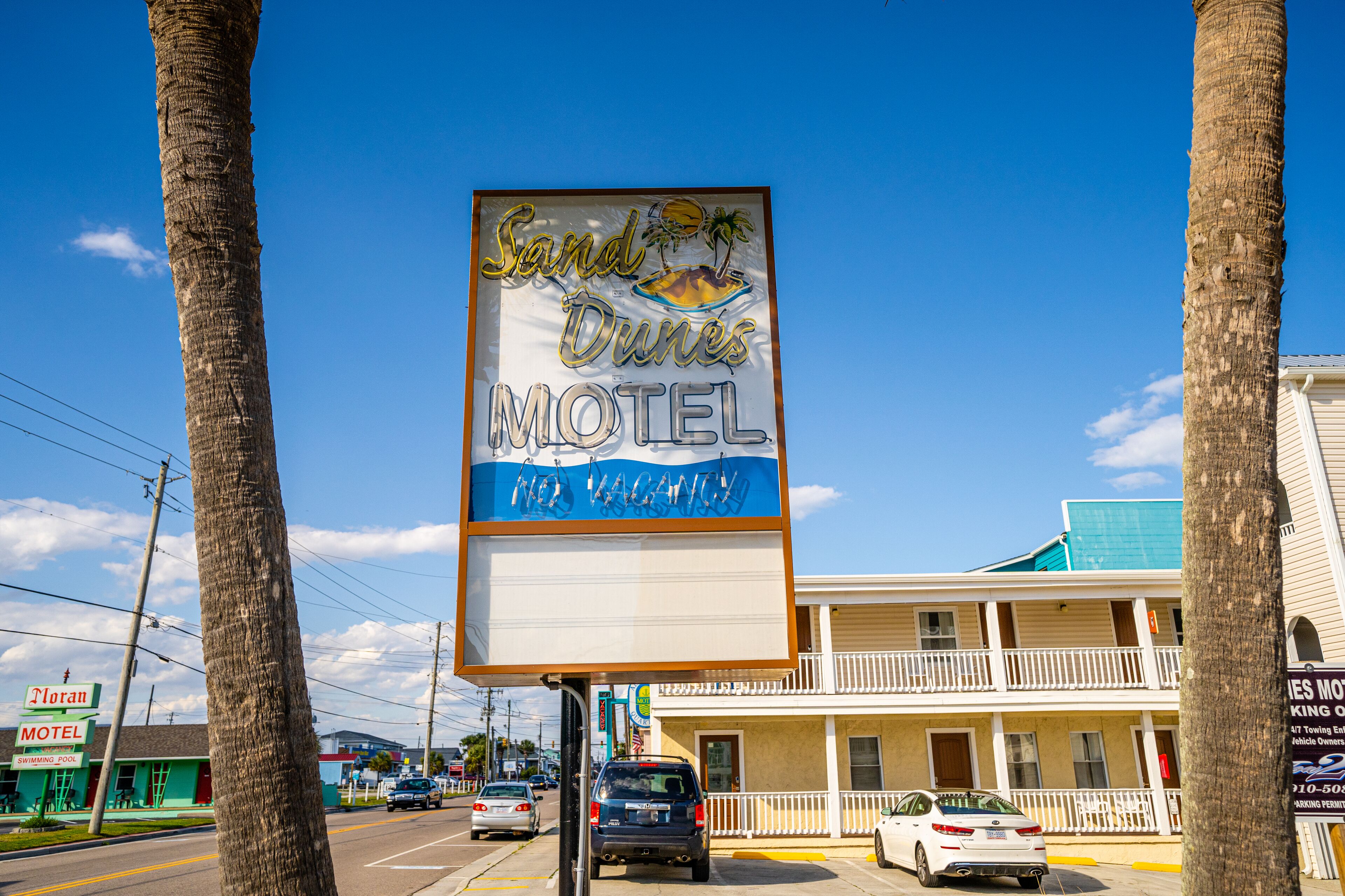 Une enseigne de motel illustrée d’une plage et d’un palmier, un bâtiment à deux étages doté d’un balcon et un parking rempli de voitures.
