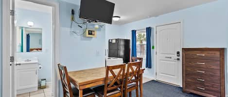 A compact room with a wooden dining table and chairs, a flat-screen TV mounted on the wall, a refrigerator, and a wooden cabinet.
