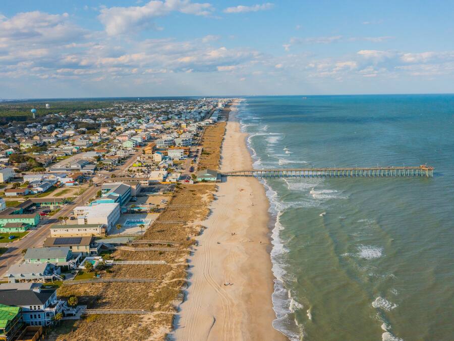 An aerial view of a coastal town with a long beach, a pier extending into the water, and residential buildings along the shore.