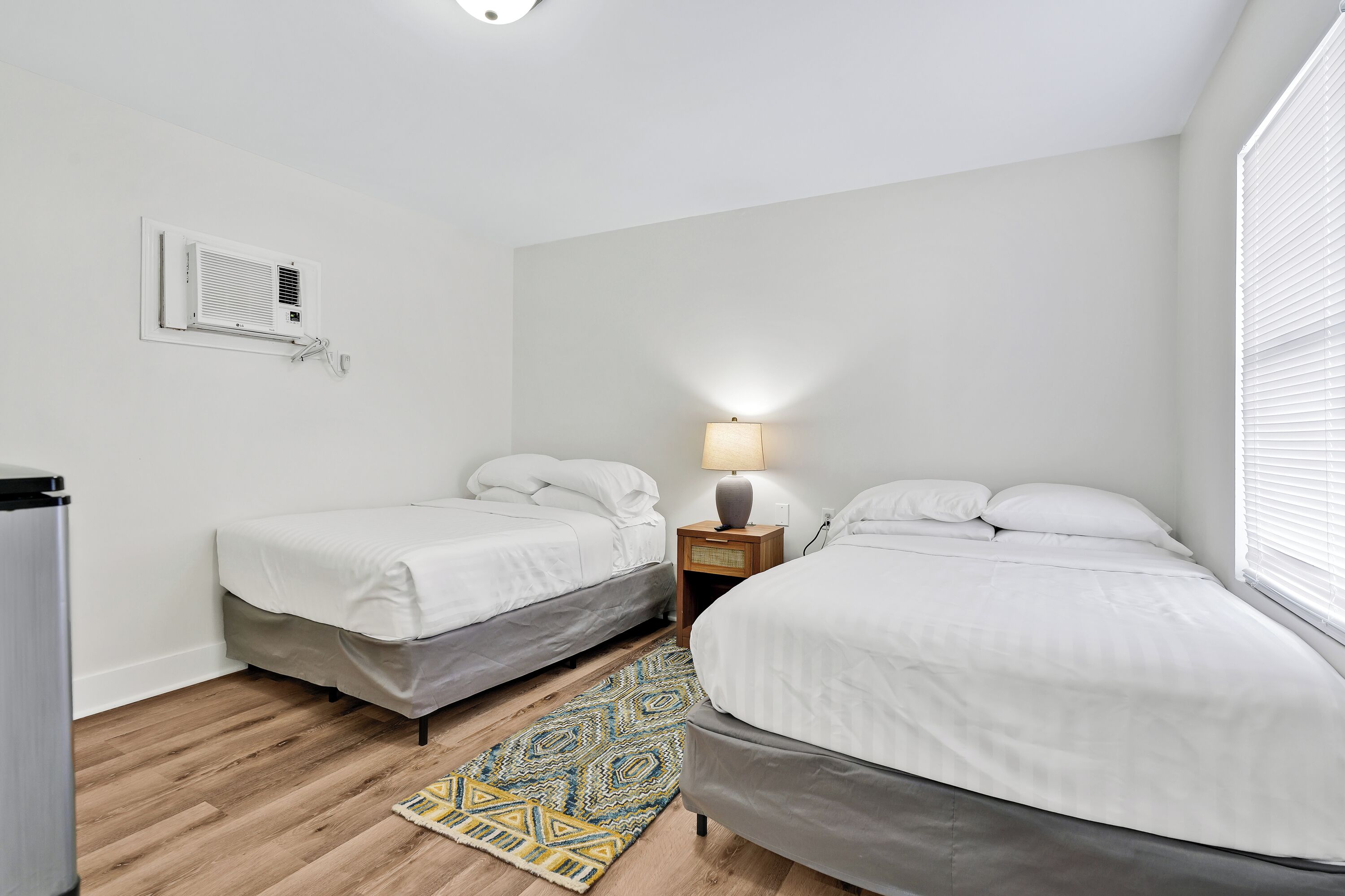 A bedroom with two single beds, a bedside table with a lamp, a wall-mounted air conditioner, and a patterned rug on the wooden floor.