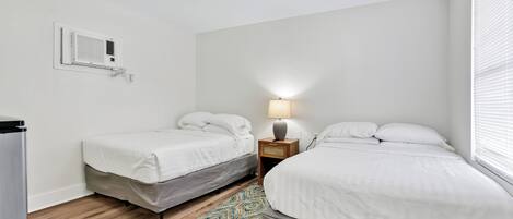 A bedroom with two single beds, a bedside table with a lamp, a wall-mounted air conditioner, and a patterned rug on the wooden floor.
