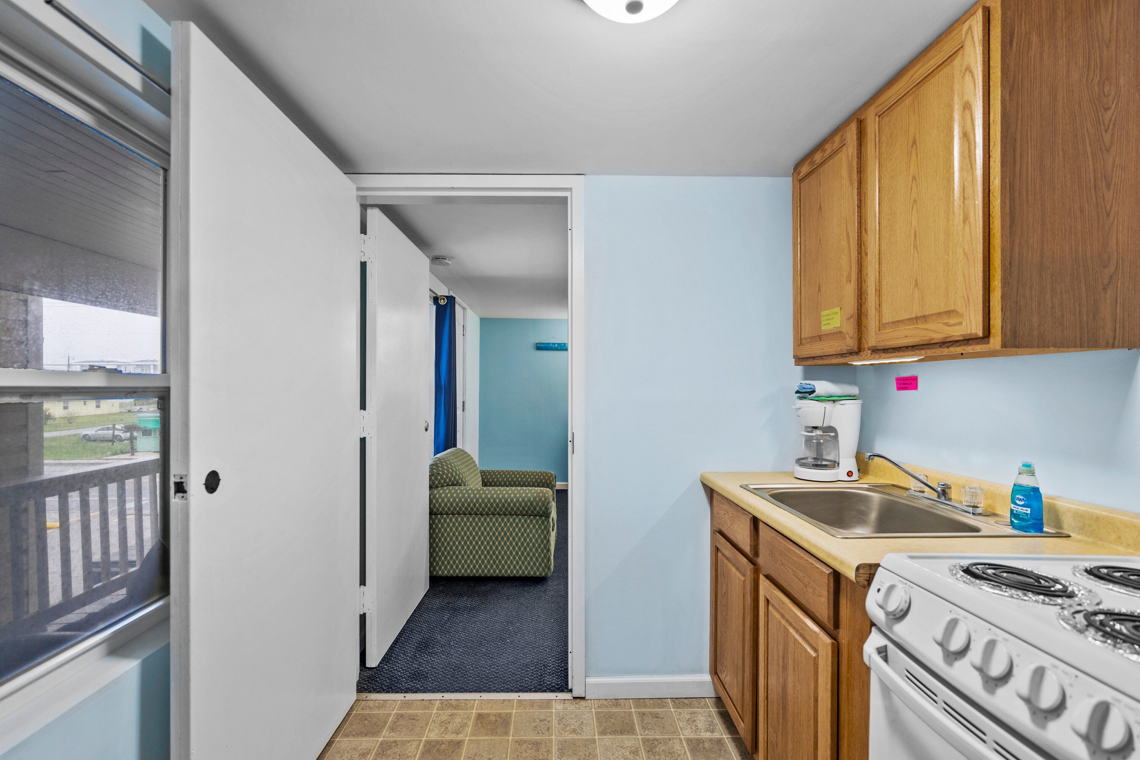 A compact kitchen with a stove, sink, and wooden cabinets. A door leads to a living area with a couch.
