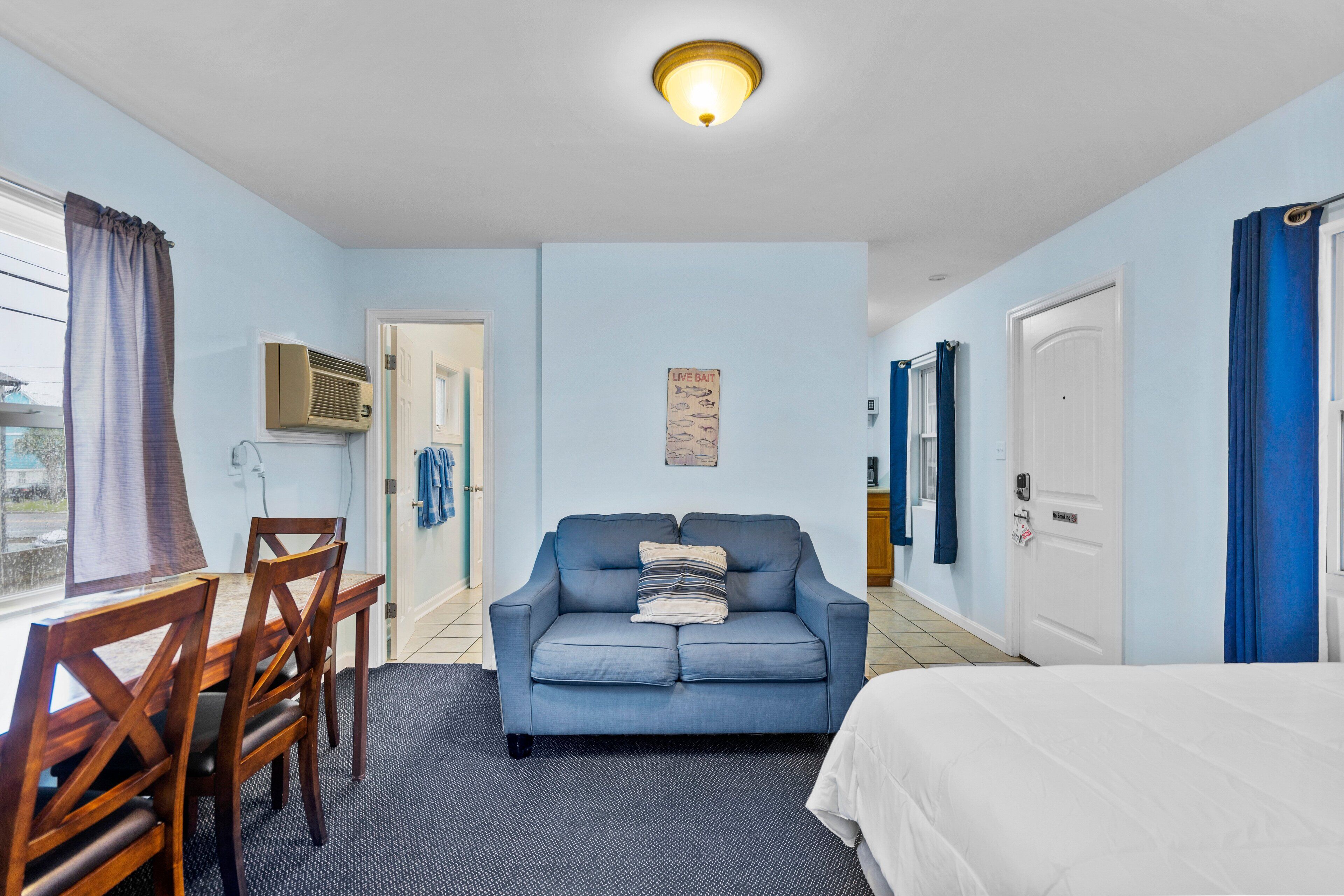 A room with a blue sofa, wooden dining table, and chairs, a bed, and a ceiling light.