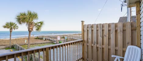 A wooden deck with a white chair, overlooking a beach with palm trees and a wooden walkway.
