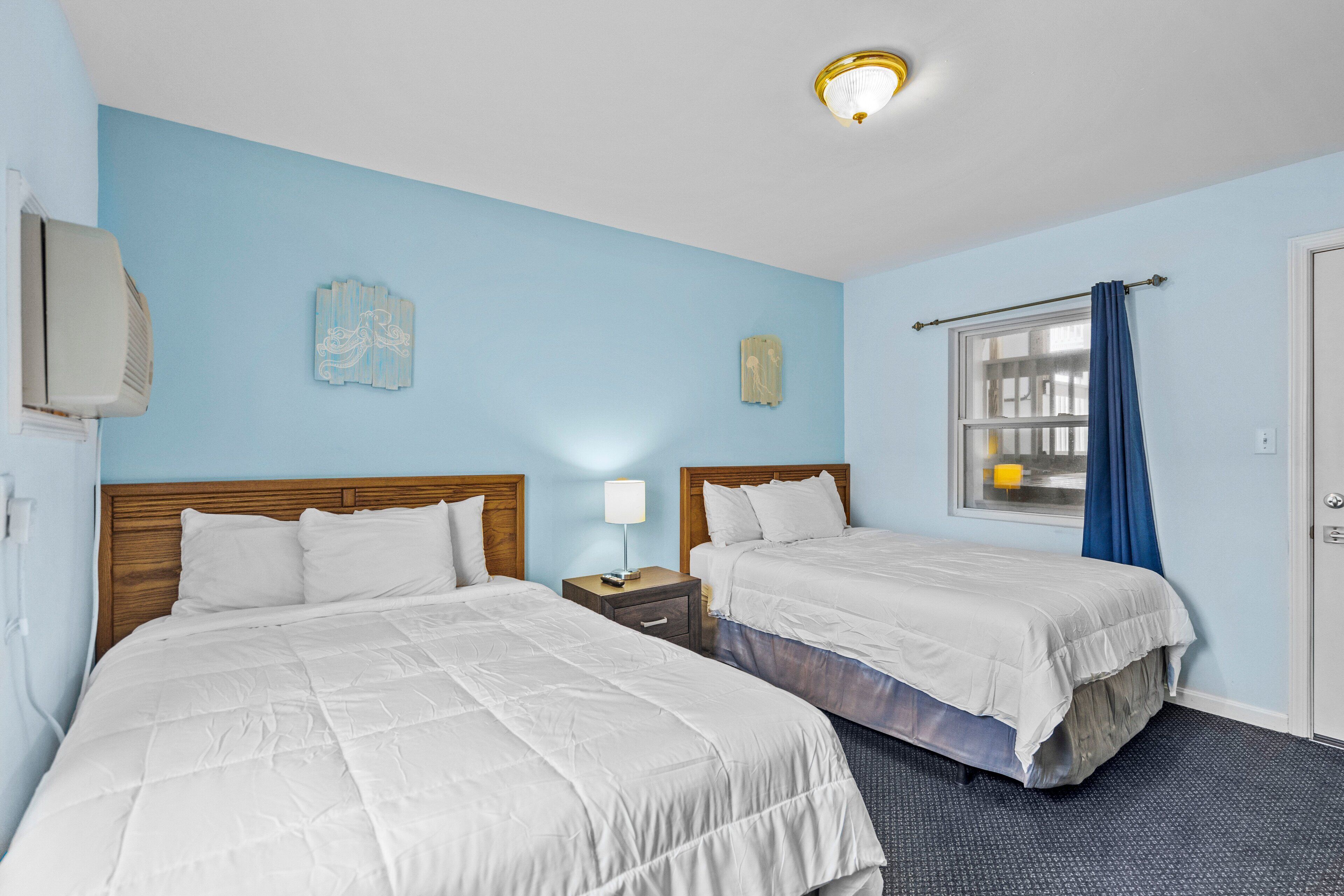 Habitación de hotel con dos camas, una mesita de noche, una lámpara y un ventanal con cortinas.