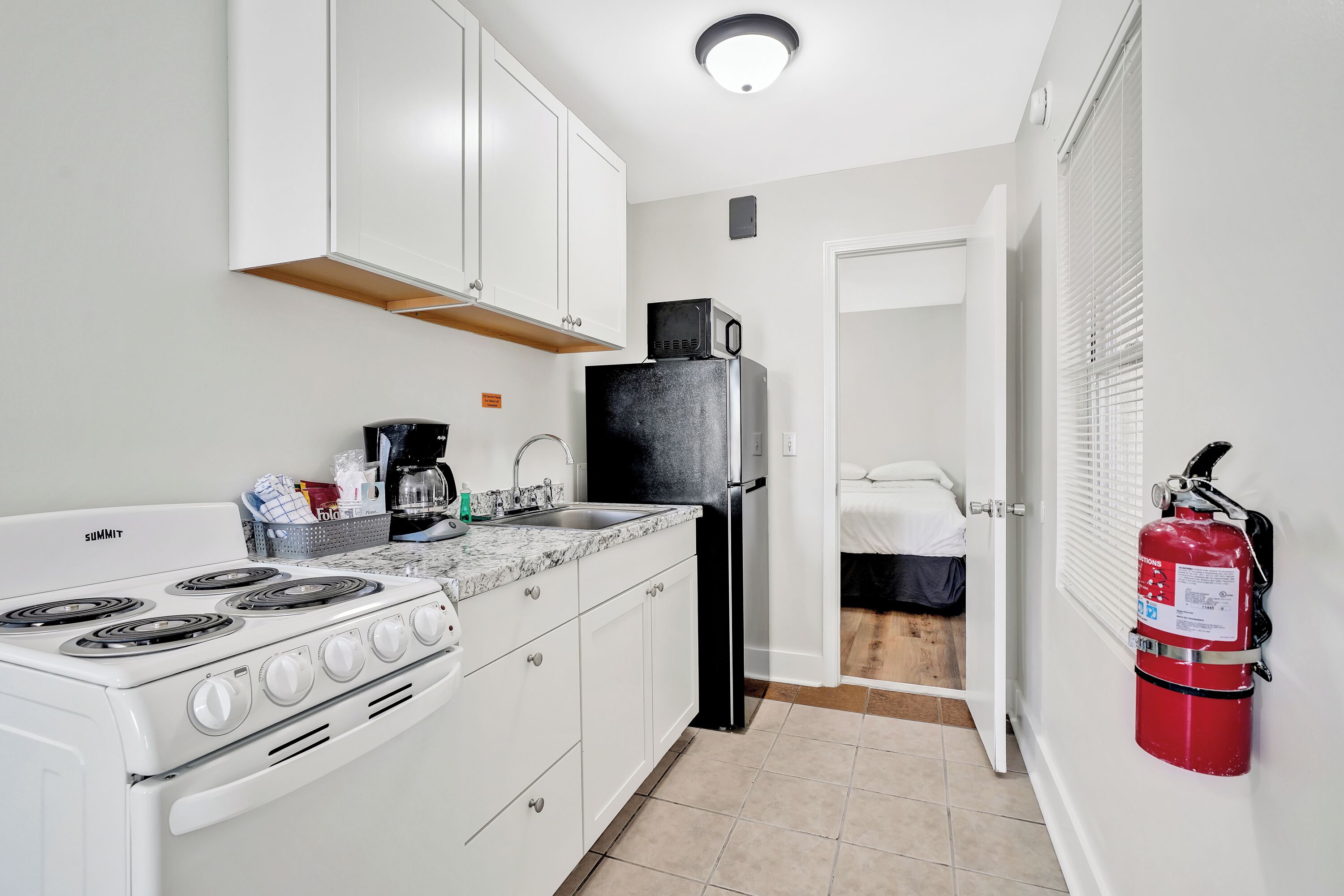 A compact kitchen with white cabinets, a stove, a sink, and a refrigerator. A fire extinguisher is mounted on the wall.