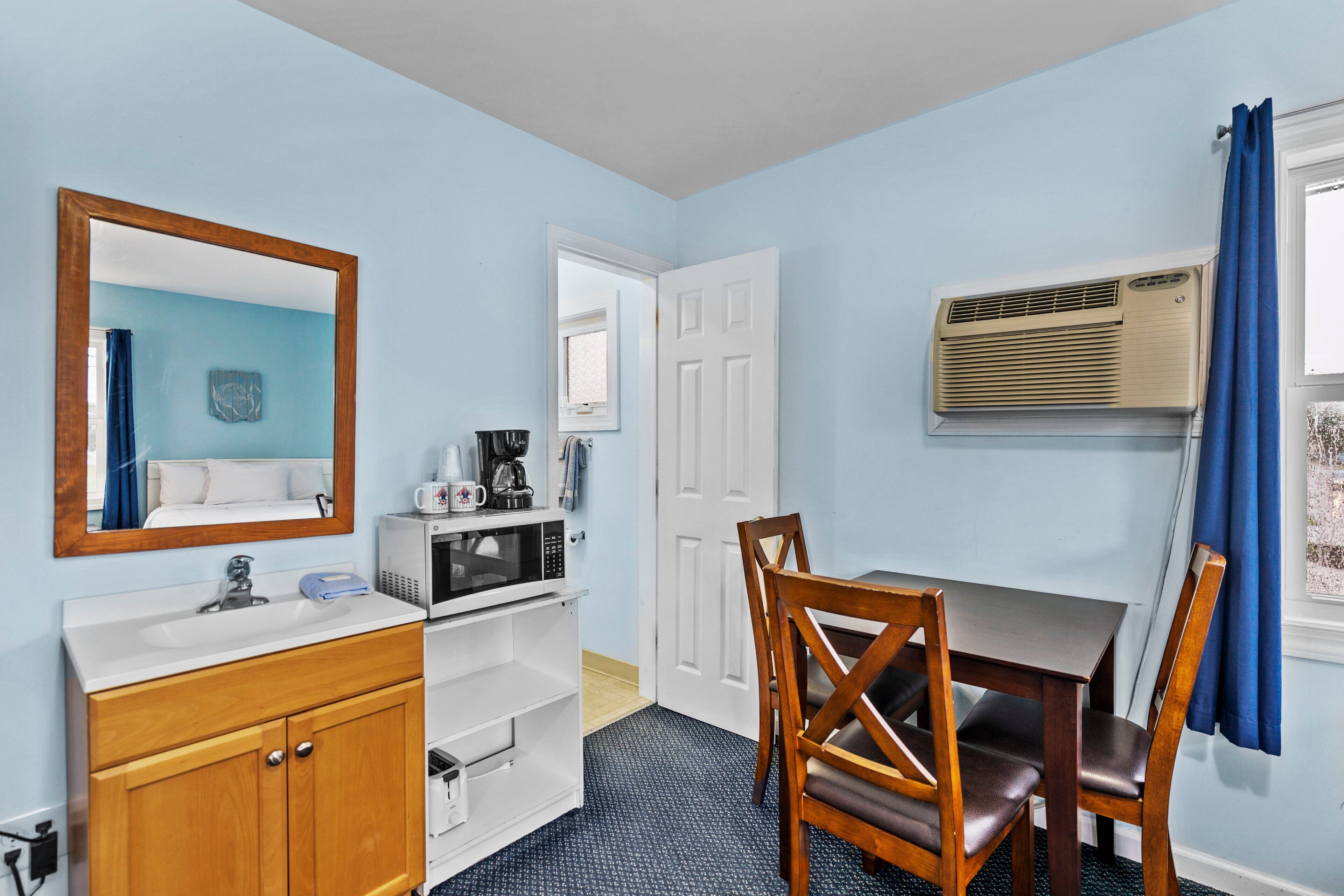 A room with a wooden cabinet, a white sink, a microwave, a coffee maker, a table with chairs, a mirror, and a window with blue curtains.