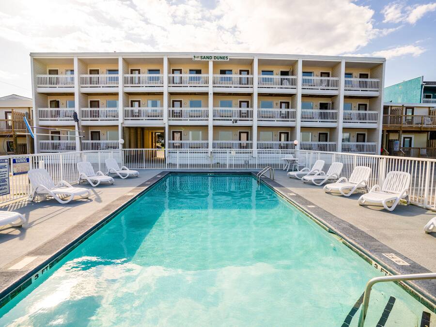 Una piscina con sillones de descanso y un edificio de varios pisos con balcones al fondo.