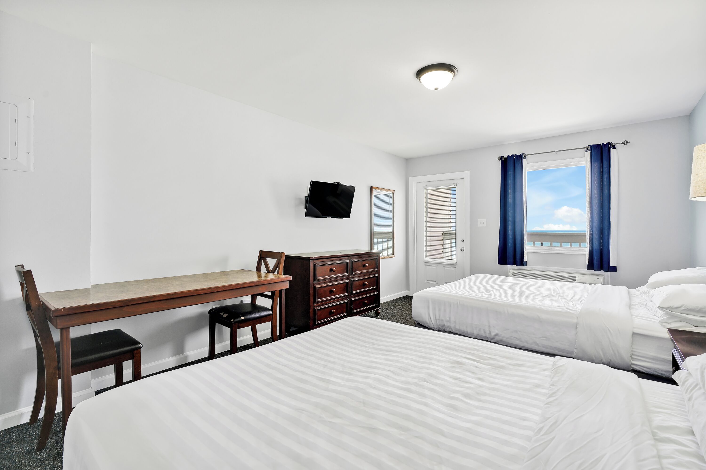 A hotel room with two beds, a desk, a dresser, a TV, and a window with curtains.