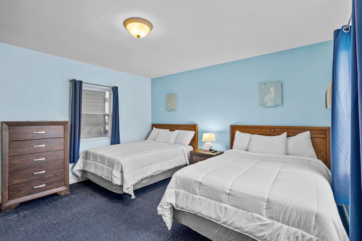 Quarto de hotel com duas camas, uma cômoda, uma janela com cortinas, uma mesinha de cabeceira com abajur e uma luminária no teto.