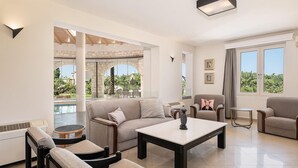 Ein geräumiges Wohnzimmer mit einem beigefarbenen Sofa, zwei Sesseln, einem Couchtisch und einem Essbereich mit Blick auf einen Pool.