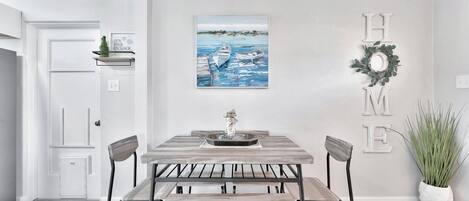 A dining area with a wooden table and chairs, a painting of boats on water, and a 'HOME' sign on the wall.