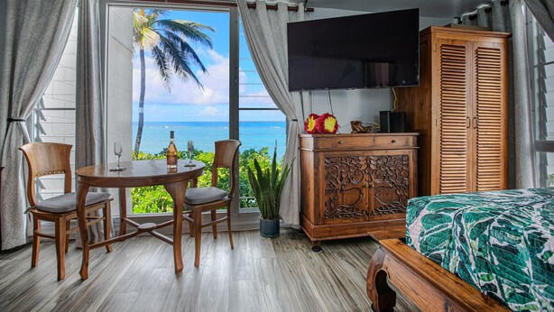 Una habitación con una cama de madera, un televisor montado en la pared, una mesa de comedor con sillas y vista al océano a través de ventanas amplias.