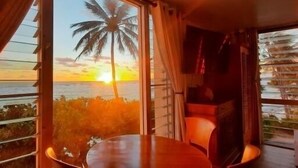 Habitación con una ventana grande que da a un atardecer y una palmera, una mesa y una silla.