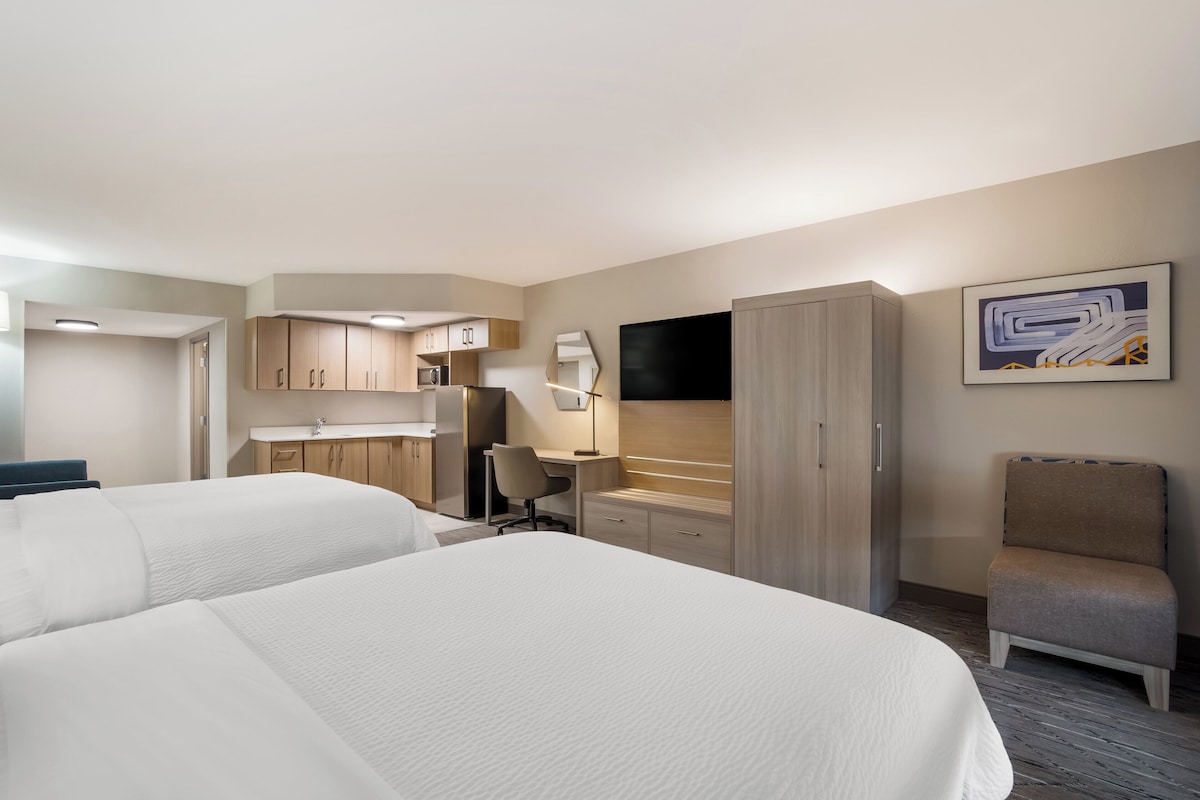 A hotel room with two beds, a desk, a chair, a TV, and a kitchenette.