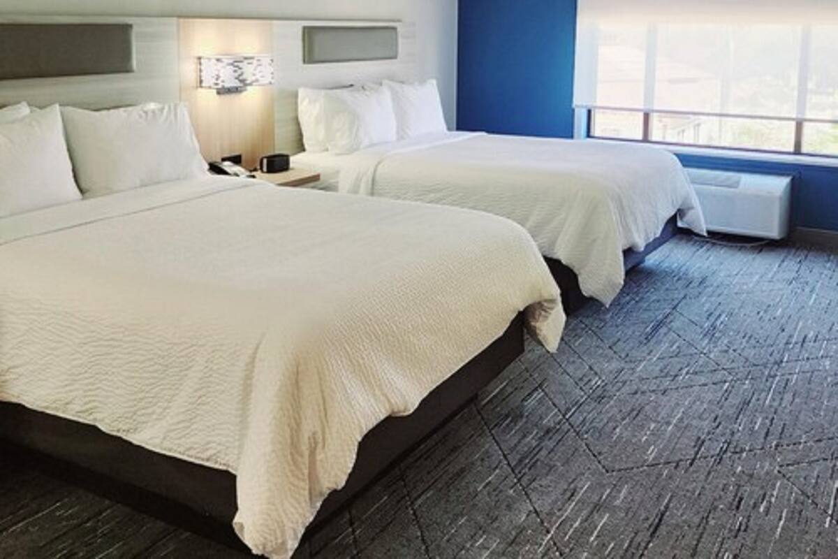 A hotel room with two beds, a nightstand, a wall-mounted TV, and a large window.