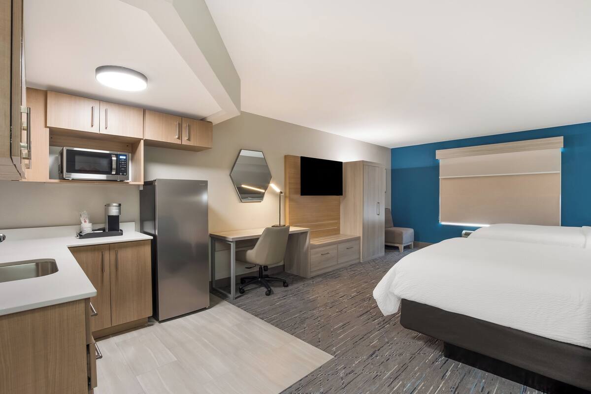 A modern hotel room with a kitchenette, a bed, a desk, and a TV.