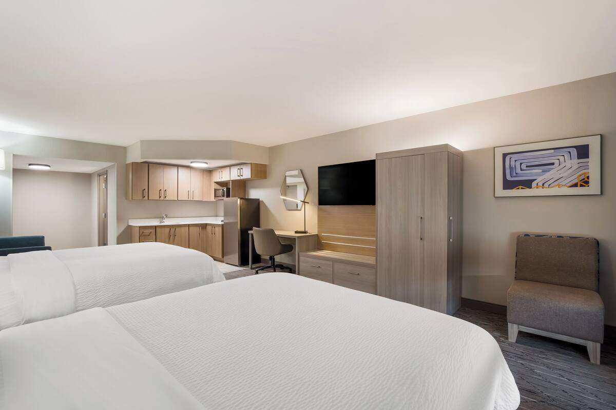 A hotel room with two beds, a desk, a chair, a TV, and a kitchenette.