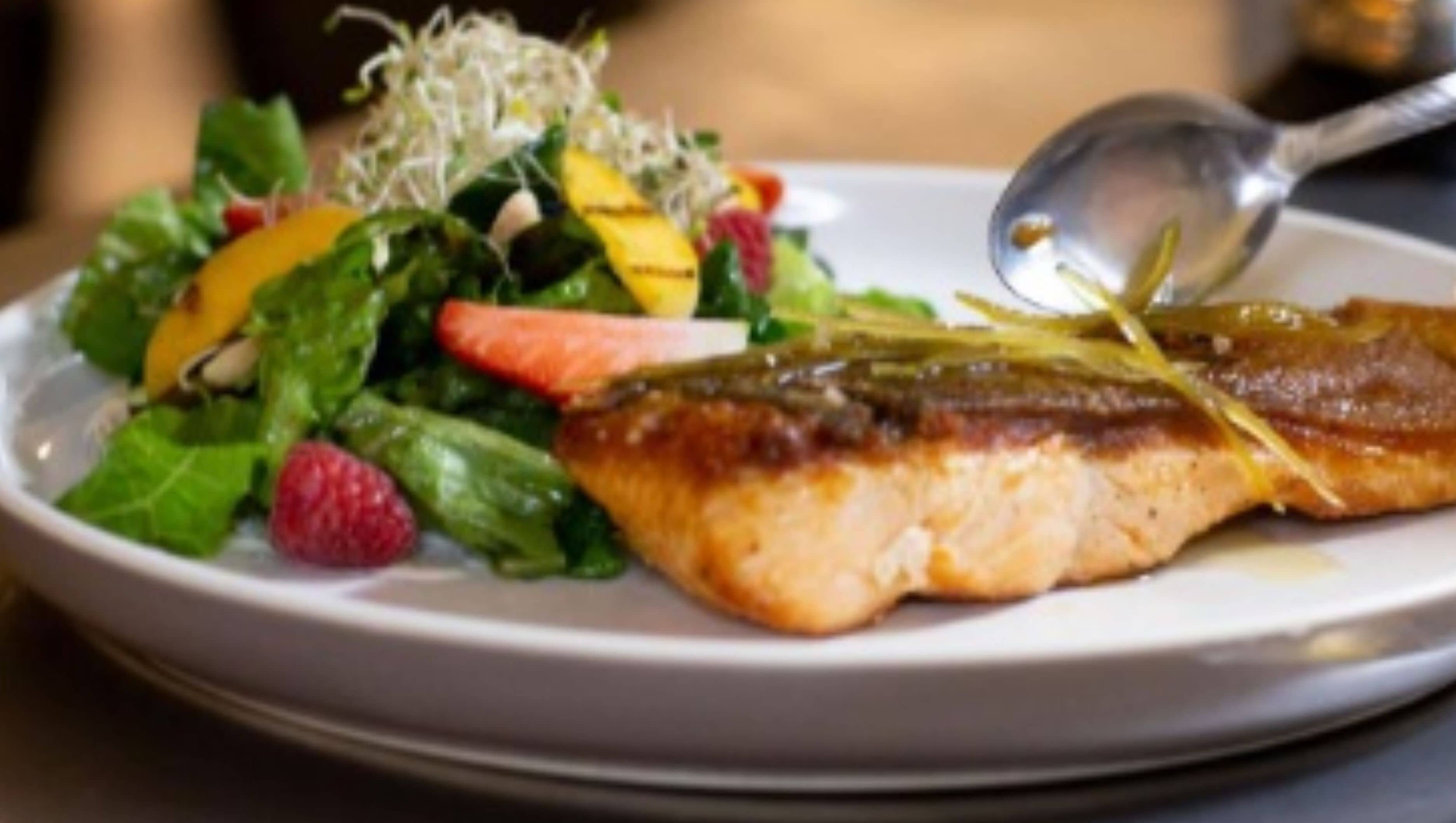 A plate with a piece of grilled fish, a salad with mixed greens and fruits, and a spoon.
