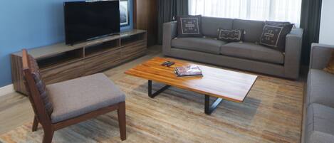 Een moderne woonkamer met een flatscreen televisie, een grijze bank met opvallende kussens, een houten salontafel en een grijze fauteuil.