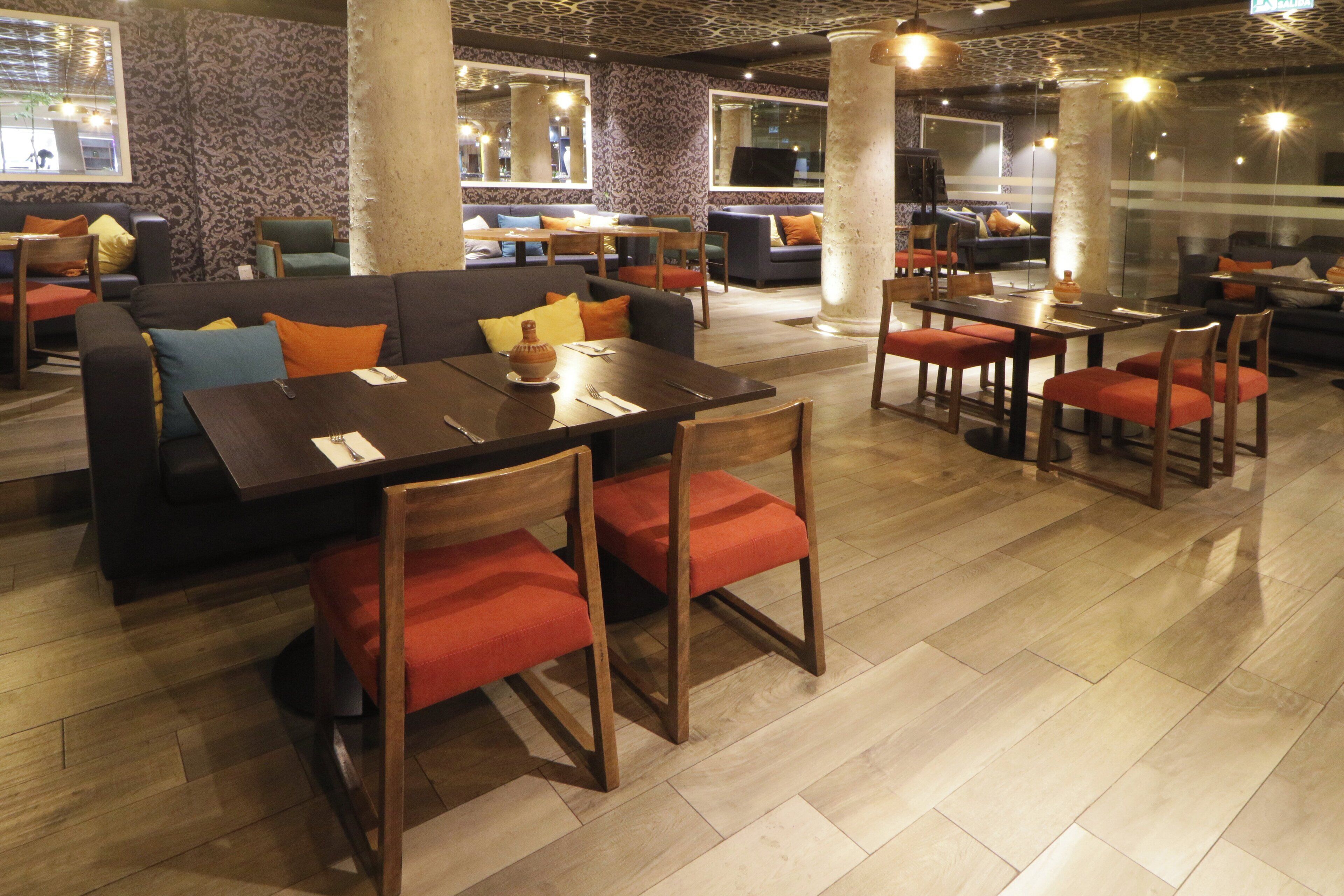 Ein Restaurant mit Holztischen und -stühlen, einer Couch mit Kissen und Wandtapete mit Muster.
