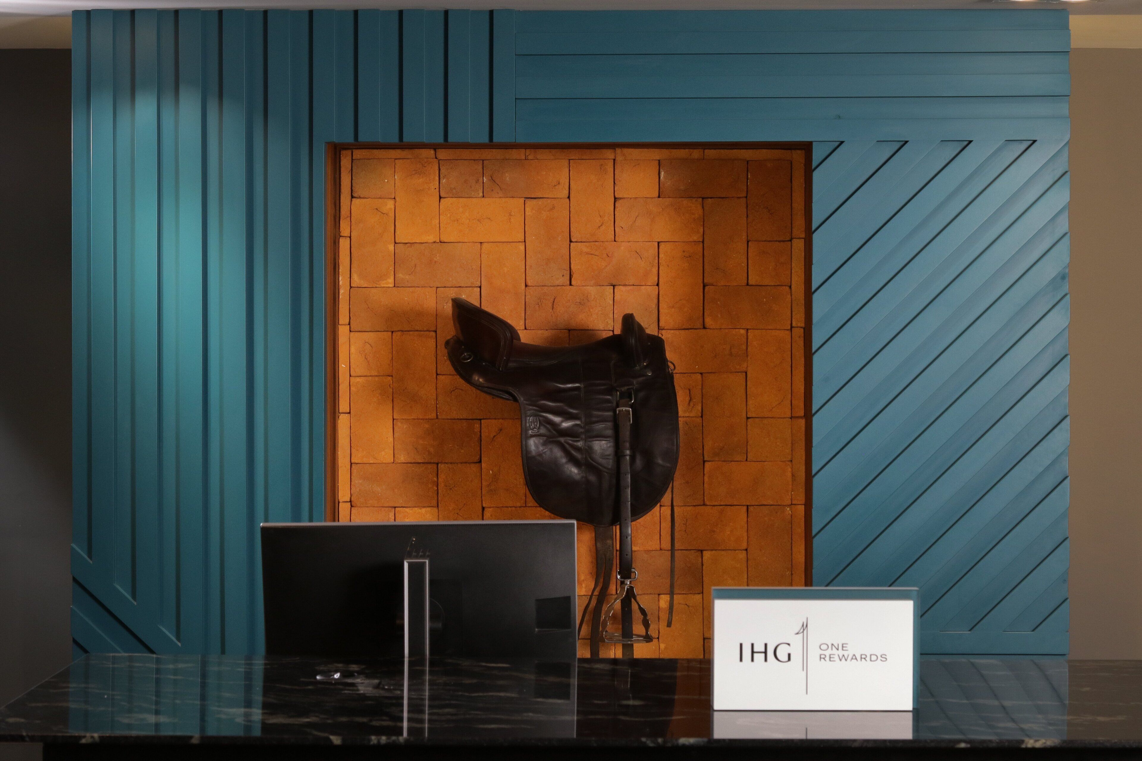 A reception desk with a laptop, a sign reading "IHG ONE REWARDS", and a wooden door with a brick wall behind it.