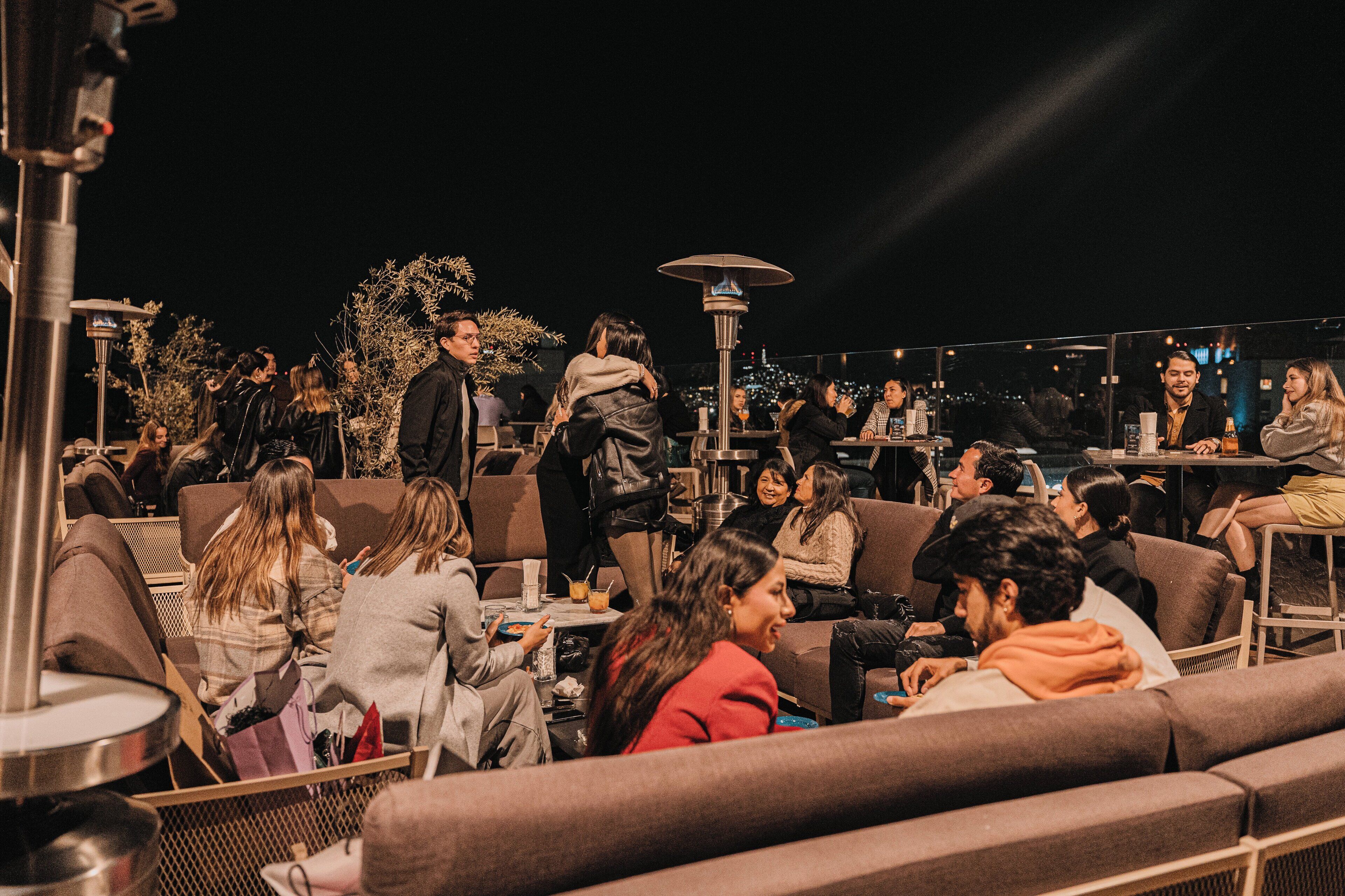 An outdoor seating area with multiple tables and chairs, surrounded by plants and a city view.