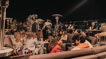 An outdoor seating area with multiple tables and chairs, surrounded by plants and a city view.