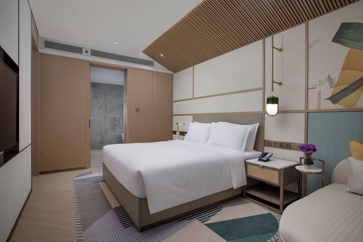 A modern hotel room with a large bed, bedside tables, a nightstand, and a wall-mounted lamp.