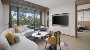Habitación de hotel moderna con una amplia televisión de pantalla plana, un sofá, una mesa de comedor y una silla. Cuenta con un balcón con vistas a árboles y una mesa con flores.