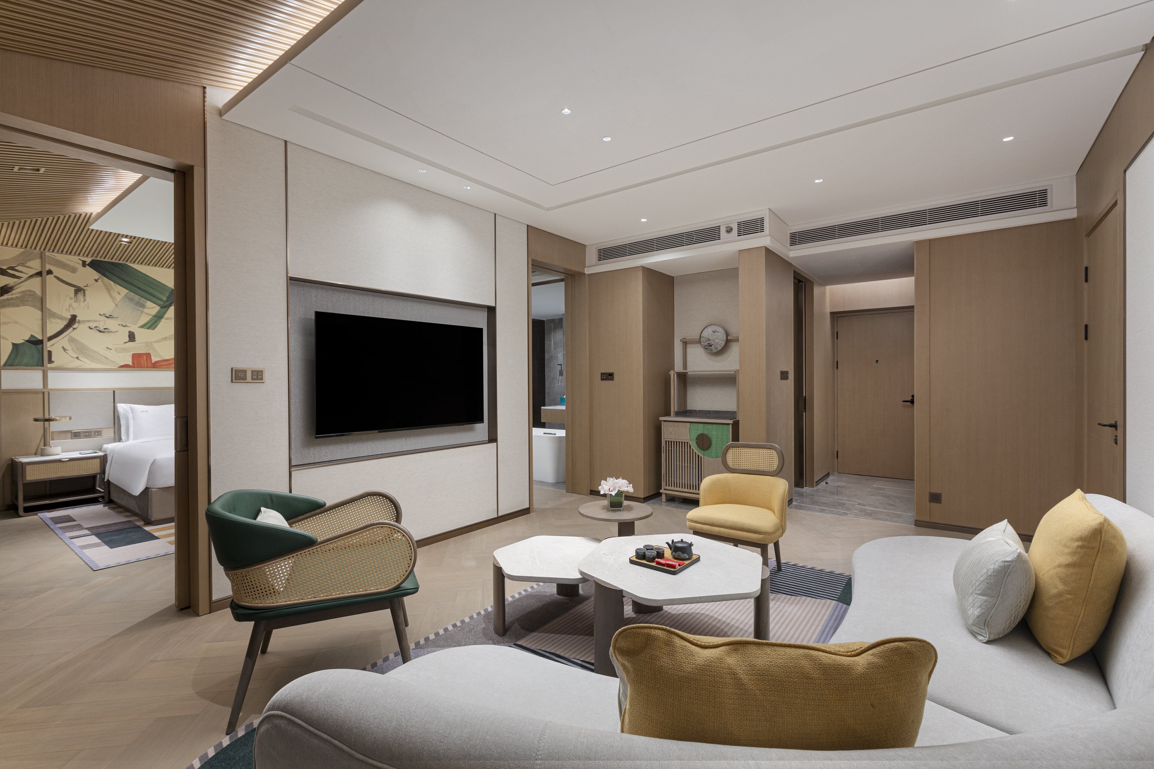 A modern hotel room with a large flat-screen TV, a sofa, a dining table, and a small kitchen area.