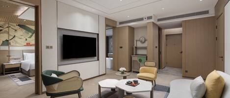 A modern hotel room with a large flat-screen TV, a sofa, a dining table, and a small kitchen area.