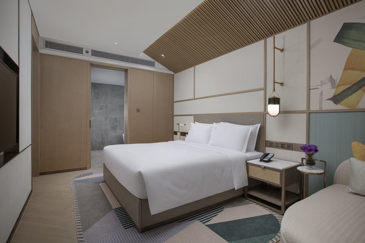 A modern hotel room with a large bed, bedside tables, a nightstand, and a wall-mounted lamp.