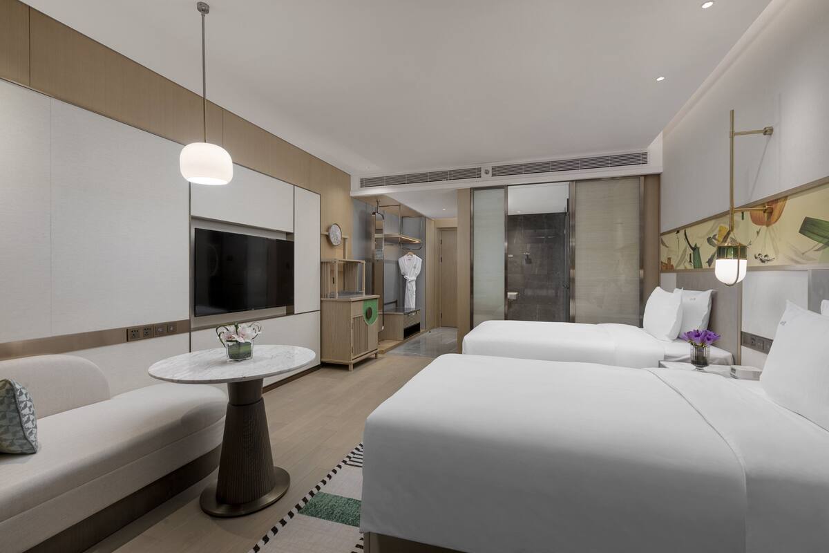 A modern hotel room with a sofa, two beds, a TV, a small table, and a bathroom with a shower and toilet.