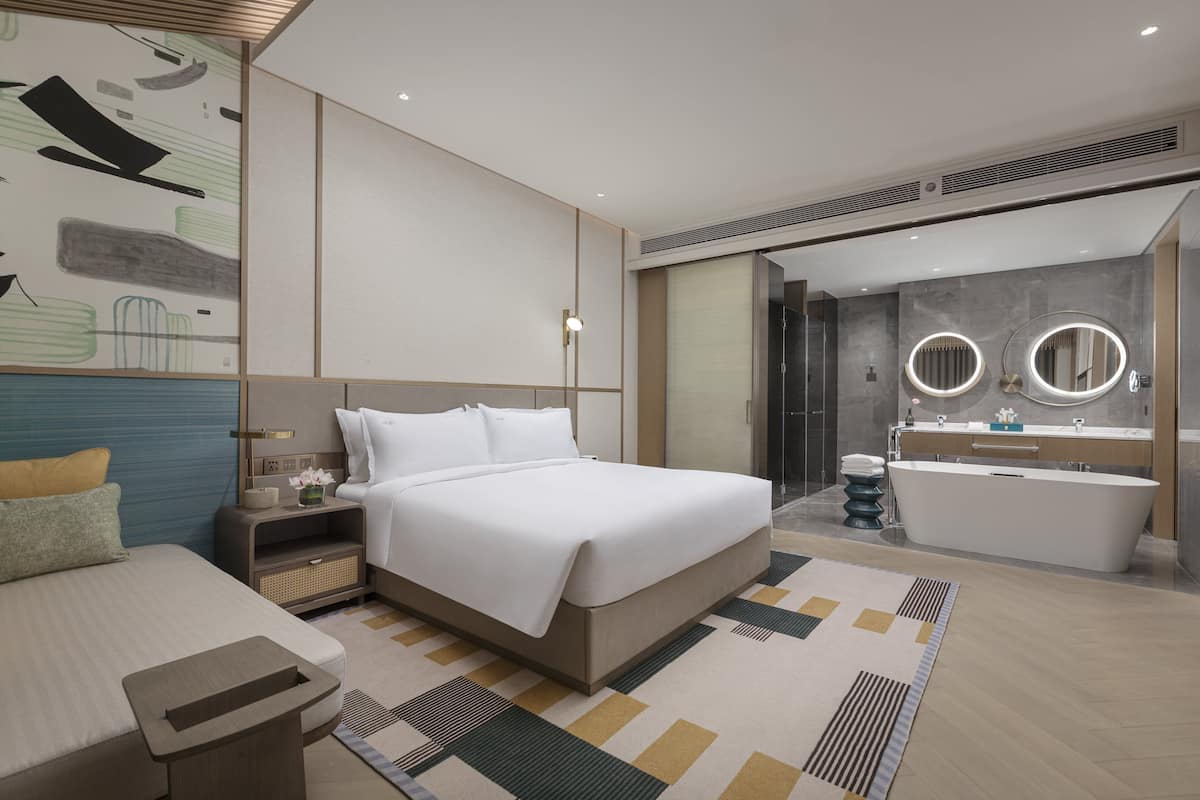 A modern hotel room with a large bed, bedside table, and a bathroom with a bathtub and two round mirrors.