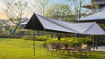 A tented outdoor dining area with a table and chairs, surrounded by a grassy lawn and trees.