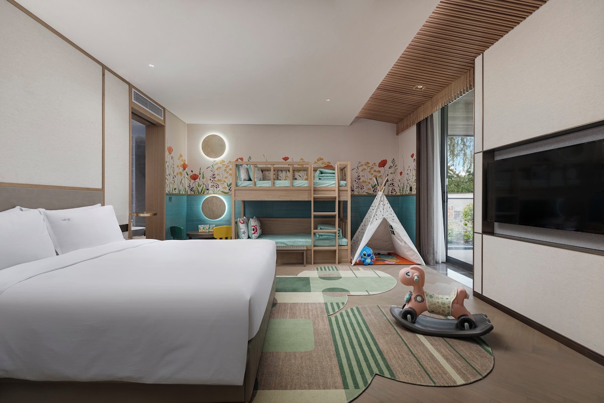A modern hotel room with a large bed, a bunk bed, a TV, and a small play area with toys.