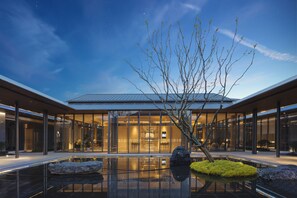 A modern building with a glass facade, a reflective pool, and a tree in the foreground. - Holiday Inn Deqing Xinshi Ancient Town by IHG (Huzhou)