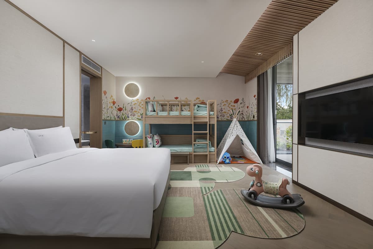 A modern hotel room with a large bed, a bunk bed, a TV, and a small play area with toys.