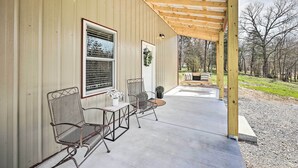A covered patio with two metal chairs and a small table, a window with blinds, and a wreath on the wall. - Eureka Springs Area Home w/ Porch, Grill! (Berryville)