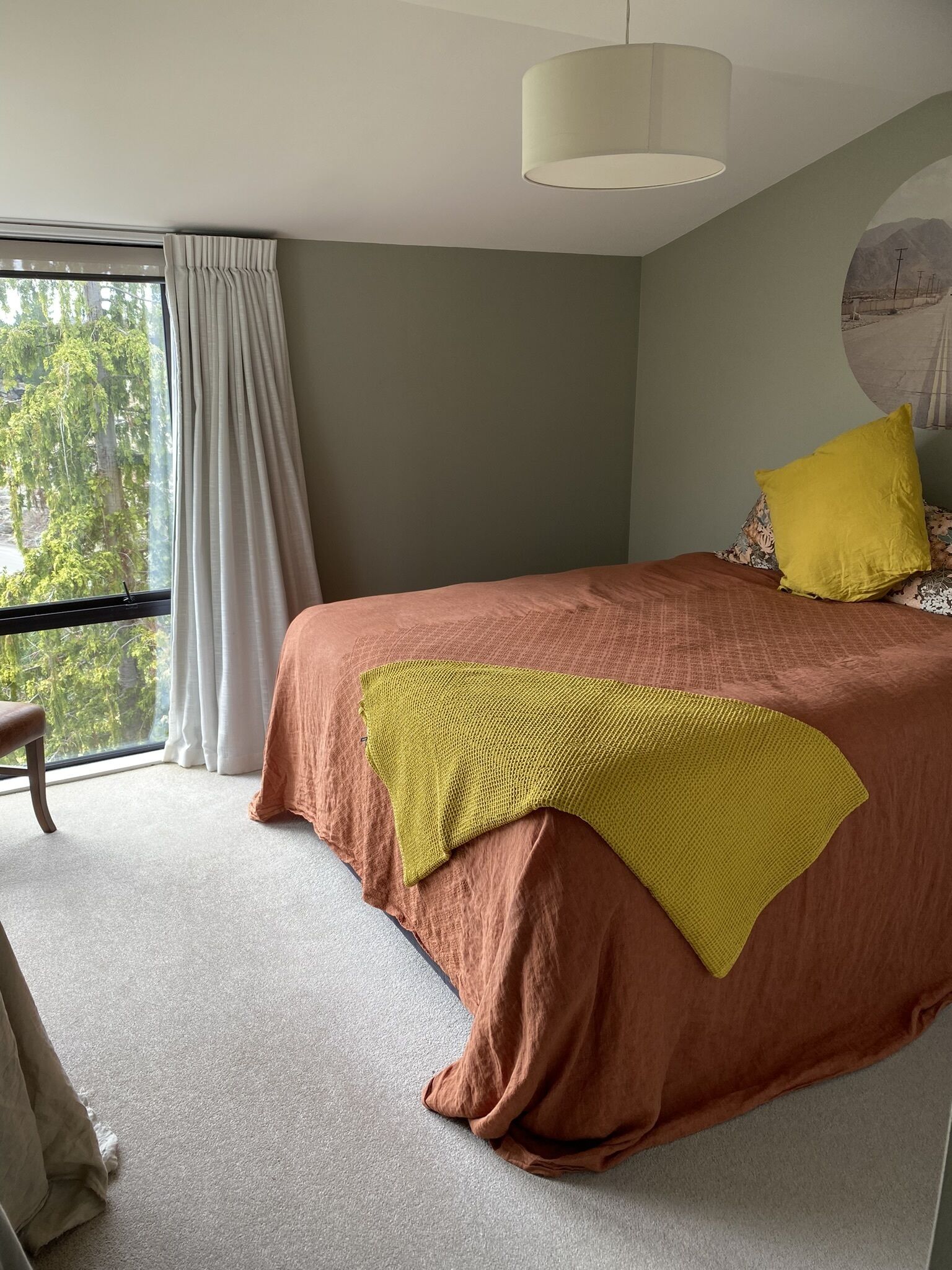 Une chambre avec un grand lit, une fenêtre avec des rideaux, un luminaire suspendu et une vue sur les arbres à l’extérieur.