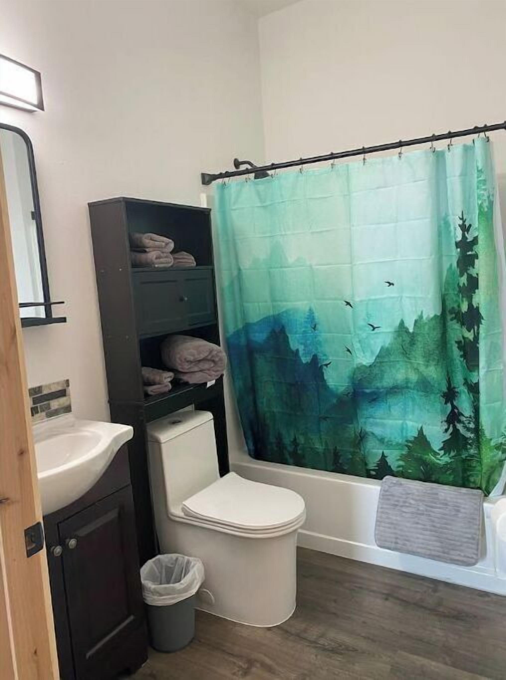 Une salle de bain avec un lavabo blanc, des toilettes, un meuble sombre, une poubelle et un rideau de douche orné d’un motif de forêt.