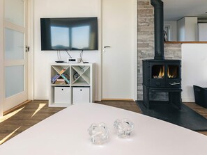 Un salon moderne doté d’un téléviseur à écran plat, d’un meuble de rangement blanc, d’un poêle à bois noir et d’une table basse blanche agrémentée de deux objets en verre.