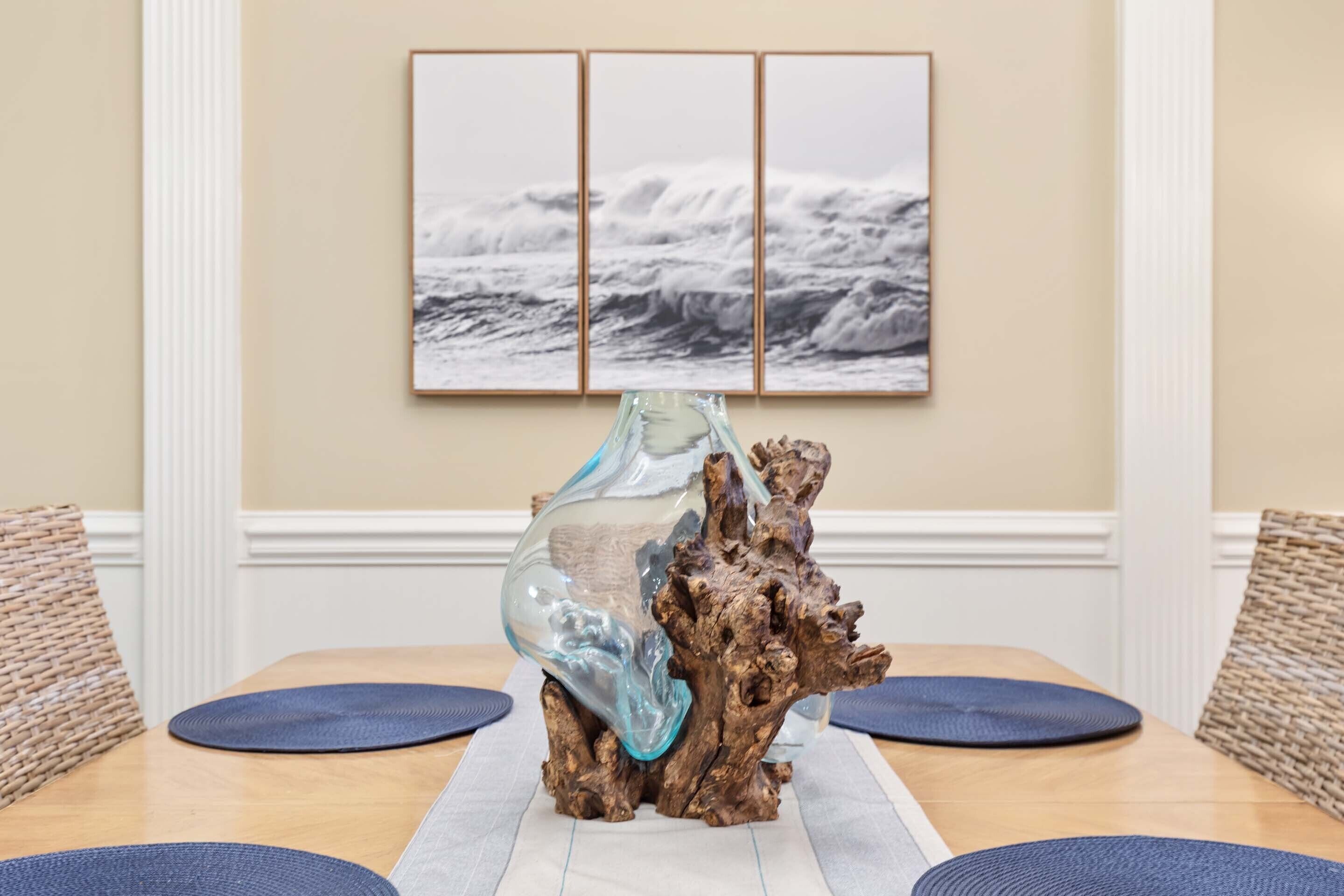 A dining table with a glass sculpture centerpiece, a framed ocean scene on the wall, and woven chairs.