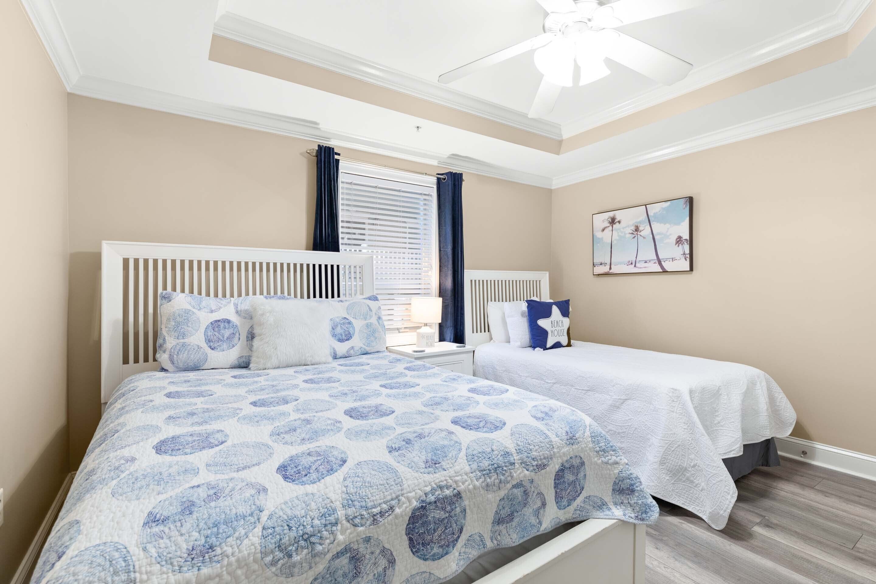 Une chambre avec deux lits, un ventilateur de plafond, une fenêtre avec des rideaux et un tableau au mur.