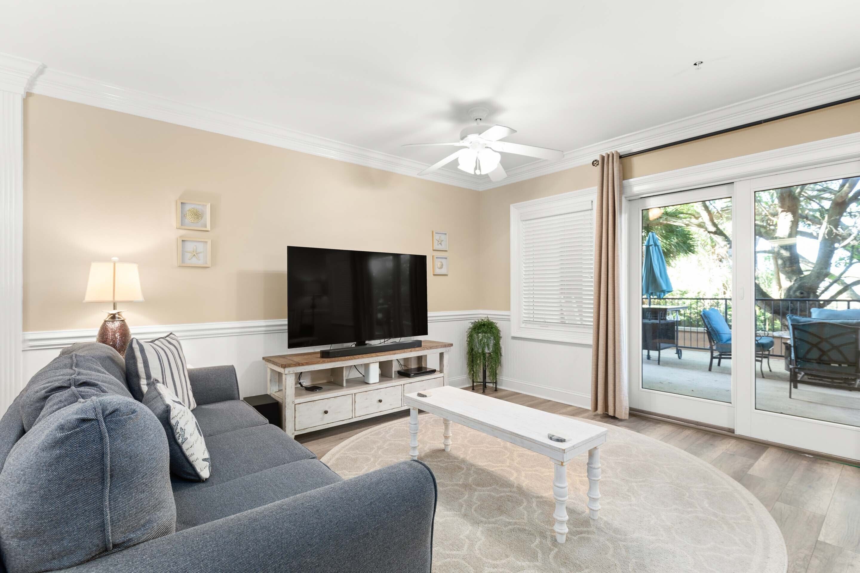 A living room with a grey sofa, a white coffee table, a flat-screen TV on a wooden stand, and a sliding glass door leading to an outdoor patio.